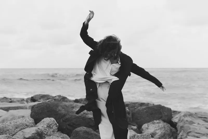 Couple dancing on rugged rocks by the sea, coats flaring, movement captured in black and white, outdoor moment of joy and togetherness, wind in hair, ocean horizon behind