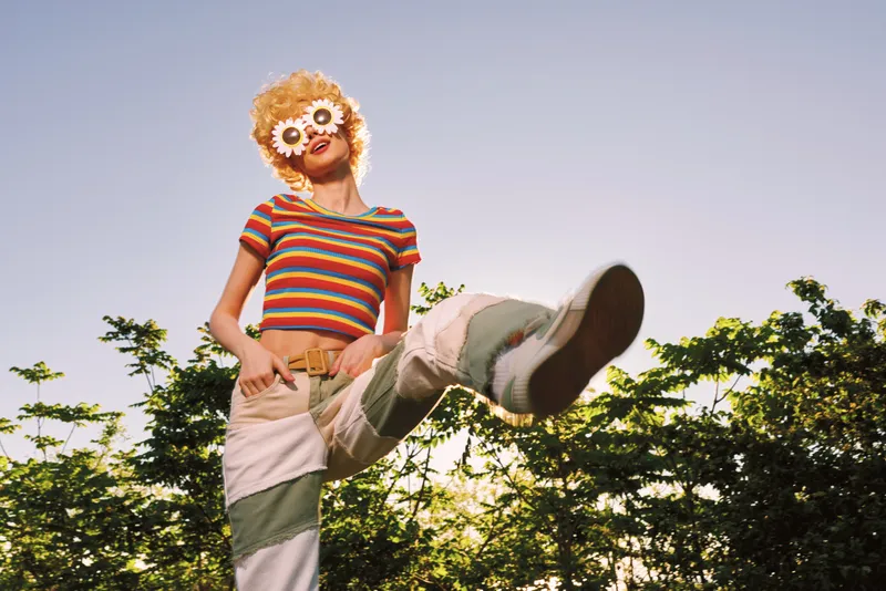 A playful woman in a striped top with a flower mask leaps outdoors, sunlit trees in the background. Retro, vibrant colors convey a carefree lifestyle moment.