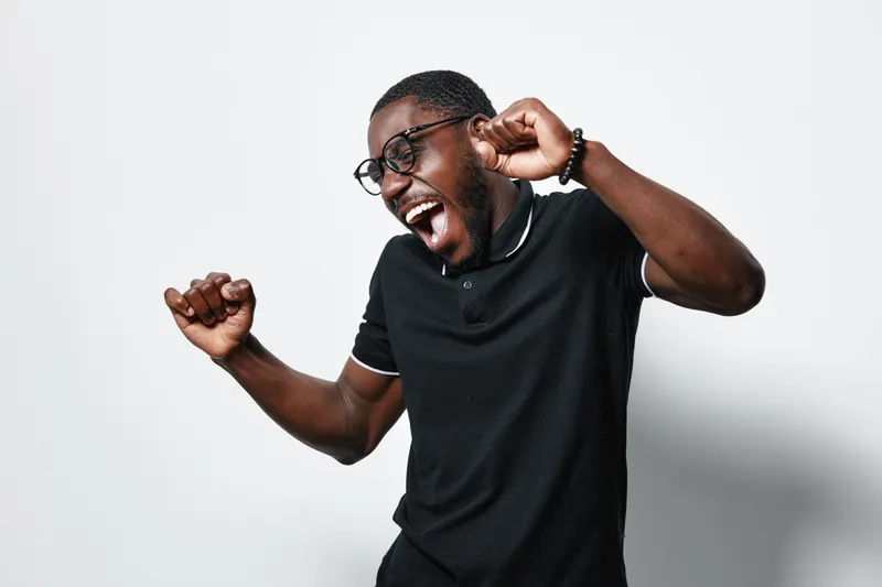 African man with dark skin and glasses wearing black polo shirt dancing and celebrating with closed eyes and raised fists on white background in studio. Joyful, happy and energetic.