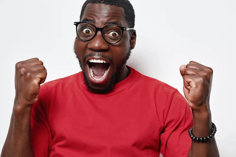 African American man with dark skin and glasses wearing red shirt celebrating success with raised fists and open mouth on white background. Excited, happy, emotional expression.