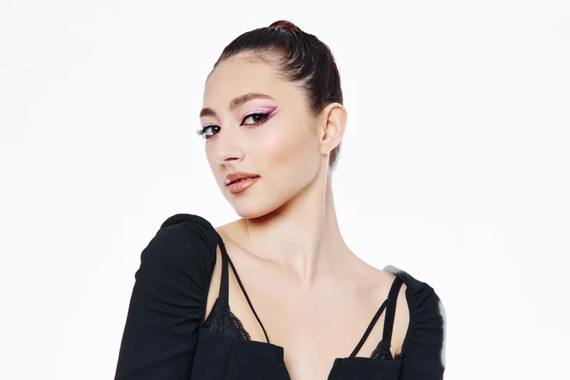 Young woman with artistic pink eye makeup poses confidently in black outfit. Studio shot on white background showcasing fashion beauty and creative makeup style.
