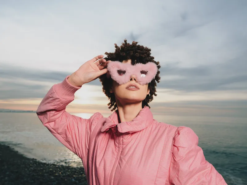 Woman wearing a pink mask and pink jacket in an outdoor seaside setting during sunset, posing confidently at the beach by the ocean and horizon with calm water