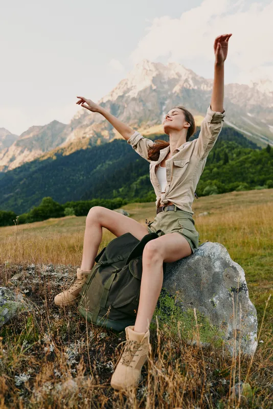 Young woman lifestyle traveler sits on rock in mountains with arms raised enjoys nature freedom and fresh air during hiking adventure in sunny day outdoors.