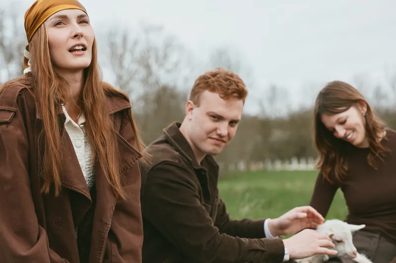 Cheerful group outdoors, goat kid presence, relaxed moment, sunlit scenery, casual outfits, candid interaction, natural colors, lush park background, wholesome lifestyle vibe