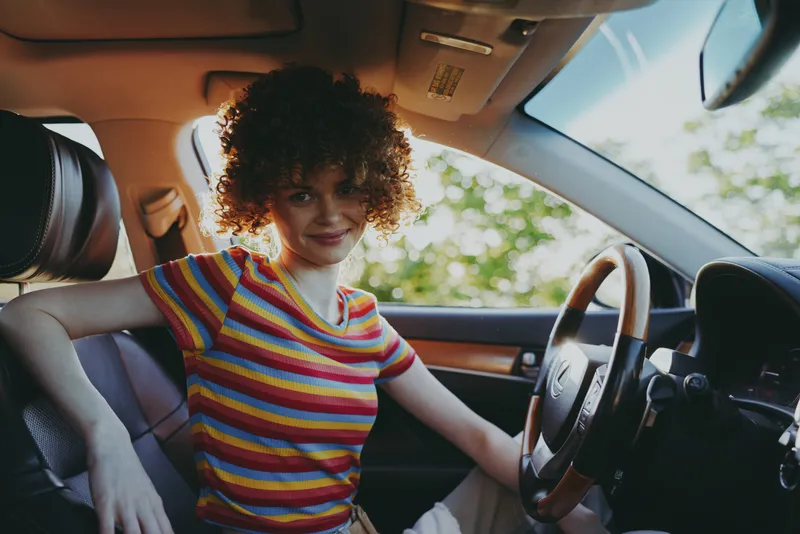 Cheerful woman in car, striped rainbow shirt, sunlit interior, relaxed posture, curly hair, warm retro vibe, natural colors, casual daytime drive moment, candid lifestyle scene