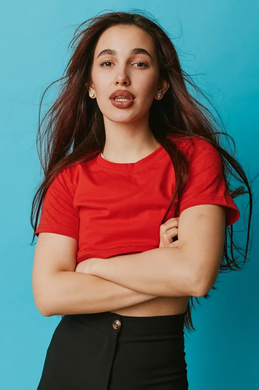 Young woman with long brown hair wearing red shirt and black pants posing with crossed arms against blue background in studio. Confident expression, casual style and natural makeup.