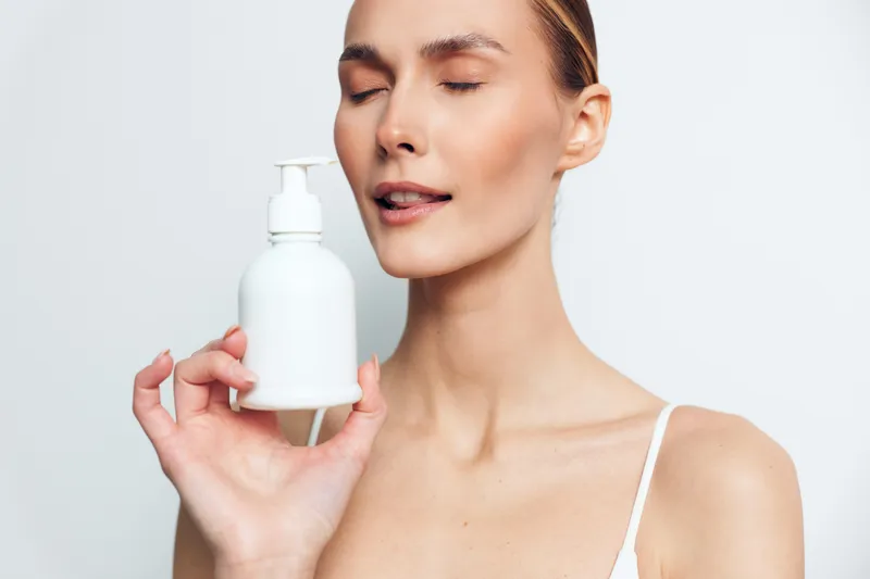Close up portrait of a serene woman holding a white skincare pump bottle, minimal studio backdrop, glowing skin, calm mood, focus on beauty routine and care