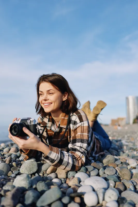 Woman photographer on a pebble beach, smiling while aiming a camera, capturing seaside scenes in bright daylight, casual plaid shirt, boots off, relaxed creative moment outdoors.