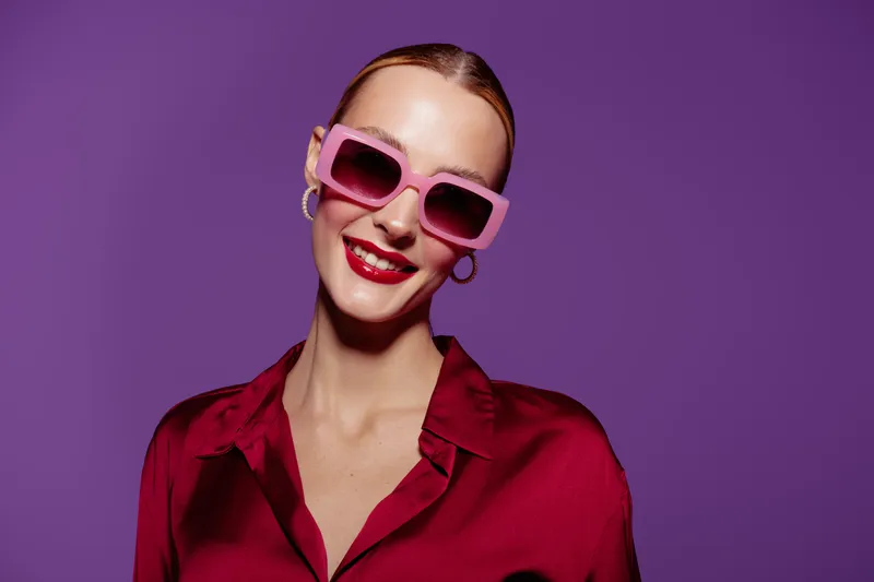 Smiling woman in red blouse wears pink sunglasses and hoop earrings against purple background. Fashion portrait with bright makeup and confident expression. Concept of style and beauty.