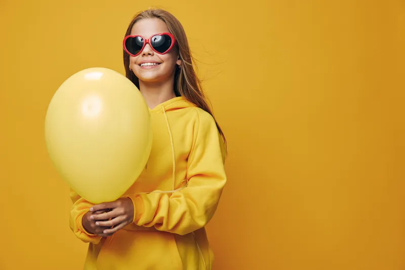 Girl with yellow balloon and wearing yellow hoodie and red heart shaped sunglasses smiling and posing on yellow background for celebration and joy.