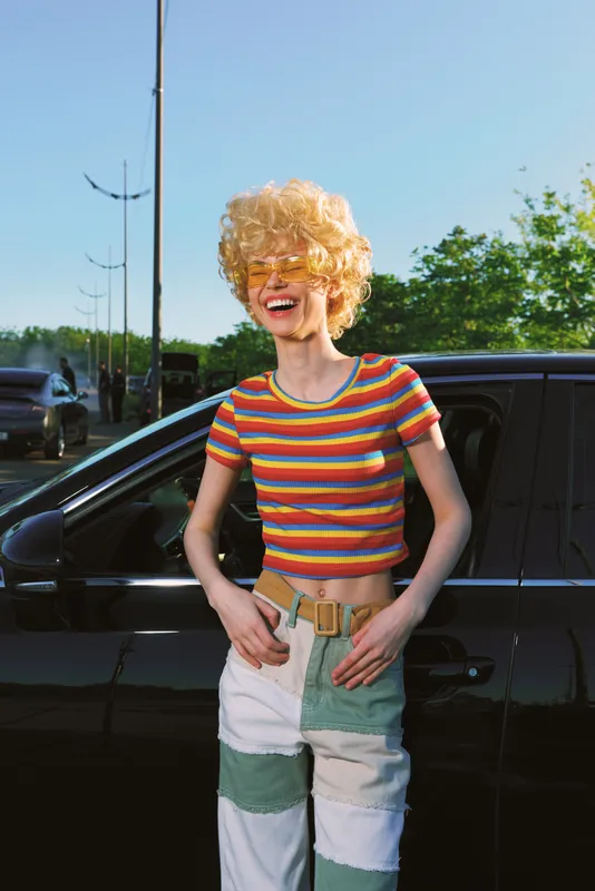 Cheerful woman outdoors in a bright striped crop top and color block jeans, smiling in sunlit breeze, retro film color vibe, casual fashion, natural greenery backdrop