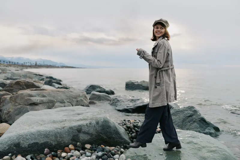 Young woman in casual clothes and cap walking on rocky beach near calm sea, smiling and looking back. Overcast sky creates soft natural light and peaceful outdoor atmosphere.