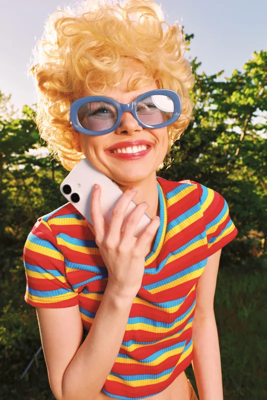 A playful blonde woman with oversized blue glasses chats on a phone in a sunny garden, wearing a colorful striped top, capturing a retro candid moment.