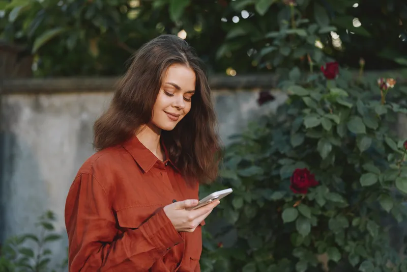 Woman in a casual orange shirt stands in a garden, looking at her smartphone with a gentle smile, natural light, greenery and flowers creating a relaxed lifestyle portrait