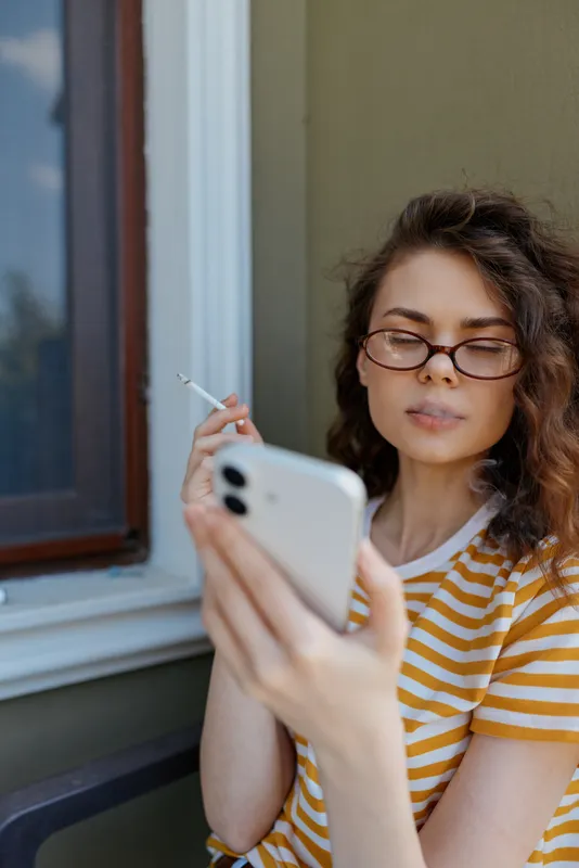 Relaxed woman at home using smartphone while smoking, casual striped tee, natural light, calm interior, focused on screen, everyday lifestyle portrait in living room