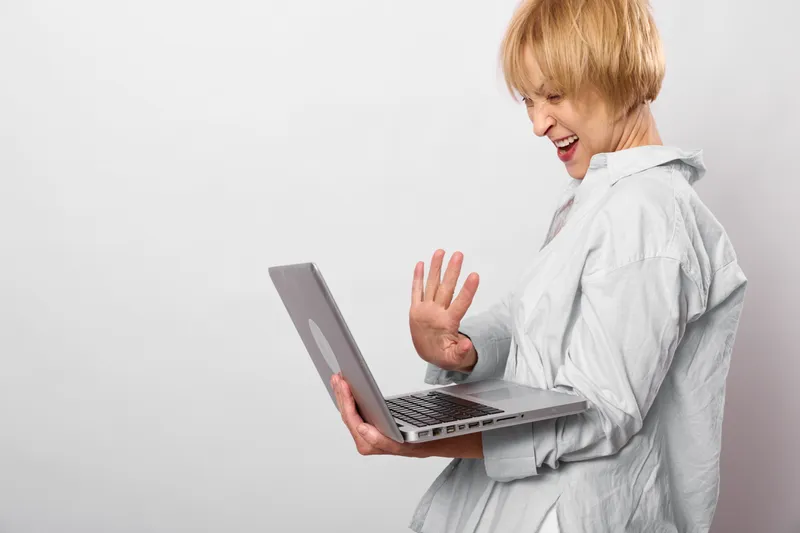 Woman laughing with laptop, hand raised, blonde portrait, white home interior, soft light, casual clothing, candid moment with open mouth and surprise gesture