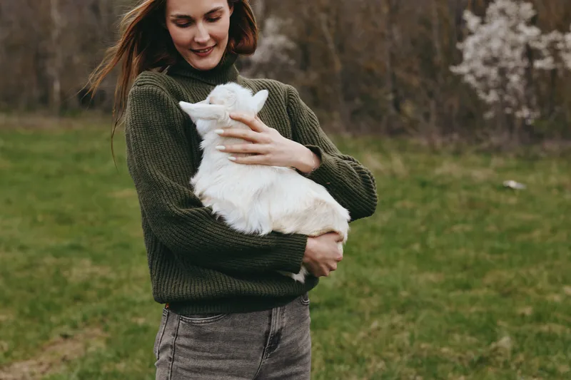 Lifestyle outdoor scene with a woman in a knitted sweater holding a white goat kid in a grassy park, calm playful mood, trees in bloom, natural rustic setting and a gentle companionship in nature.