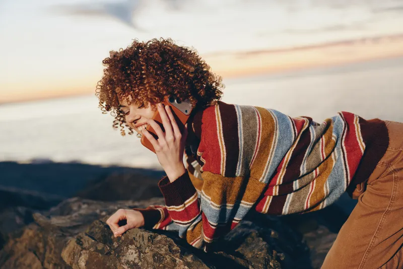 woman outdoors lifestyle curly hair striped sweater sunlight, she touches her face beside beach rocks, smiling softly, calm relaxed mood, warm breeze, natural tones in golden light today