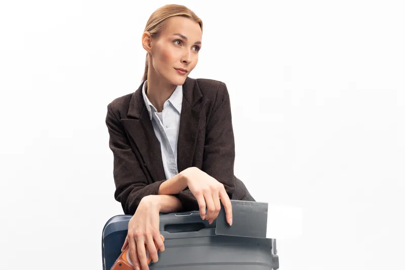 Young woman in formal jacket holding a suitcase and passport on white background. Concept of travel preparation, business trip, and vacation planning with confident expression and neat appearance.