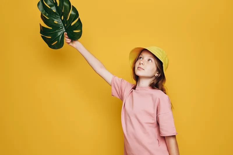 Young girl in a yellow hat and pink shirt holds a large green leaf against a yellow background. Studio shot with bright colors and natural concept themes.