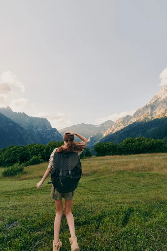 Hiking woman with backpack in a sunlit meadow, mountains in the background, exploring nature and outdoors in a vibrant travel moment