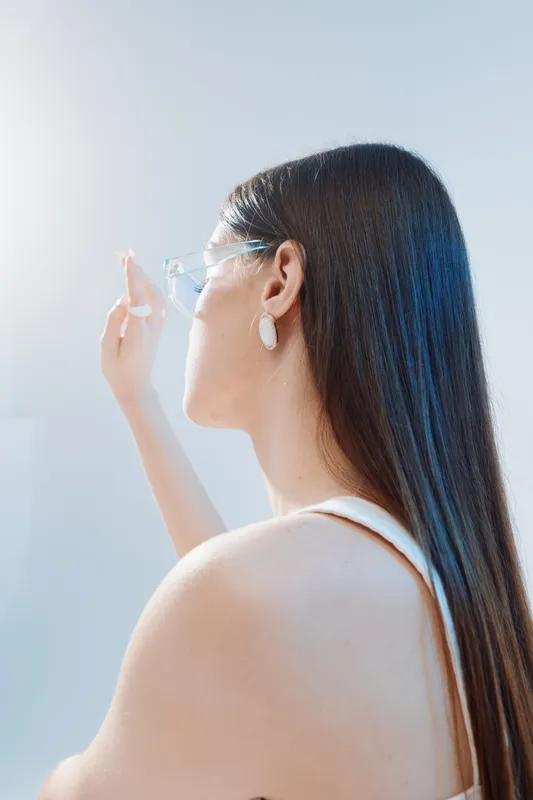 Young woman with long hair and clear glasses looks away while reaching out her hand. Soft natural light highlights her smooth skin and elegant posture. Concept of beauty fashion.