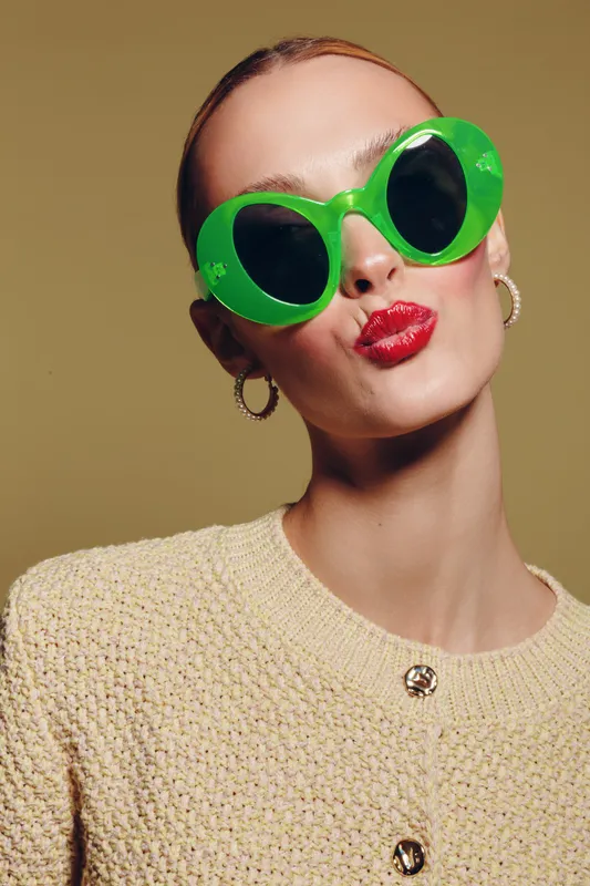 Young woman with bright red lipstick wearing large green sunglasses and beige knitted sweater poses against plain background. Concept of fashion, style, and bold accessories.