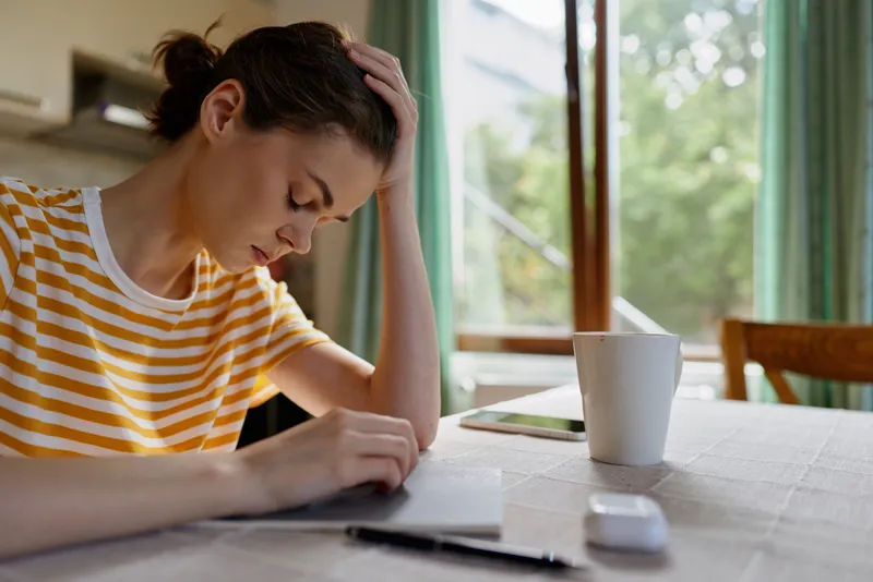 A stressed woman in a striped tee sits at a home desk, head resting on her hand while she writes on papers, with a mug beside the window, conveying fatigue and focus.
