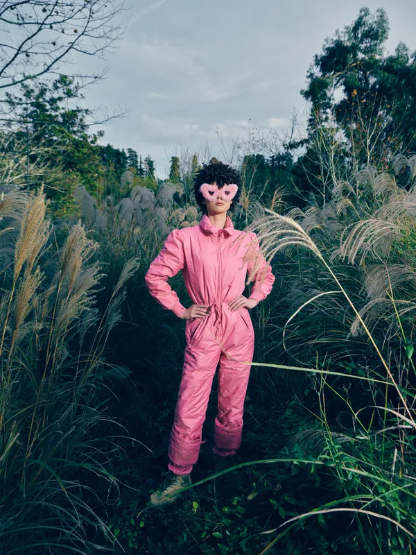 Woman in a pink jumpsuit stands confidently among tall grass, wearing a decorative mask, outdoors, bold fashion, natural light and dramatic color in a lush landscape.