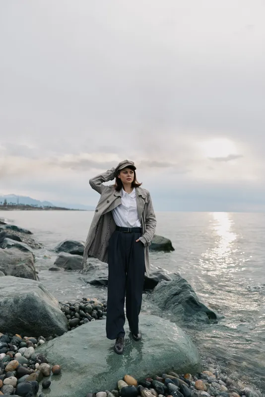 Young woman stands on rocky shore near calm sea wearing coat and cap. Overcast sky and reflective water create moody atmosphere with cool tones and natural textures.