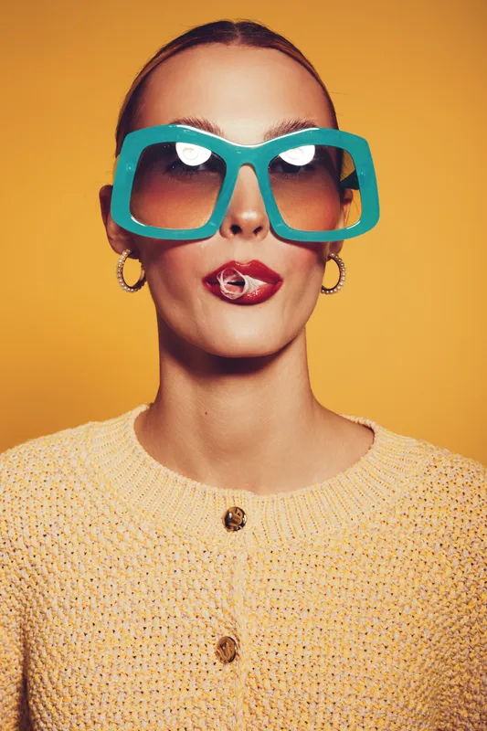 Stylish woman with oversized turquoise sunglasses and red lipstick smokes cigarette against yellow background. Fashion portrait with bold accessories and confident expression in studio lighting.