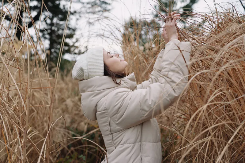 Winter outdoor scene with a woman in a light beige coat and knit hat reaching for tall grass in a quiet field, capturing a moment of calm, exploration and natural textures.