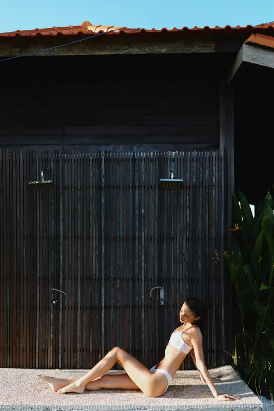 Bright outdoor scene shows a woman in a bikini relaxing by a rustic doorway in a sunlit courtyard, a casual summer lifestyle with gentle breeze, warm tones, and vibrant plants