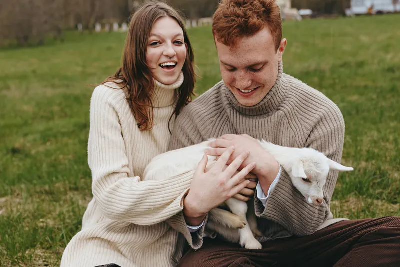 Smiling couple outdoors with a goat kid resting in their lap, wearing cozy sweaters. Natural light, warmth, affection, and countryside field vibes convey lifestyle and animal bonding.
