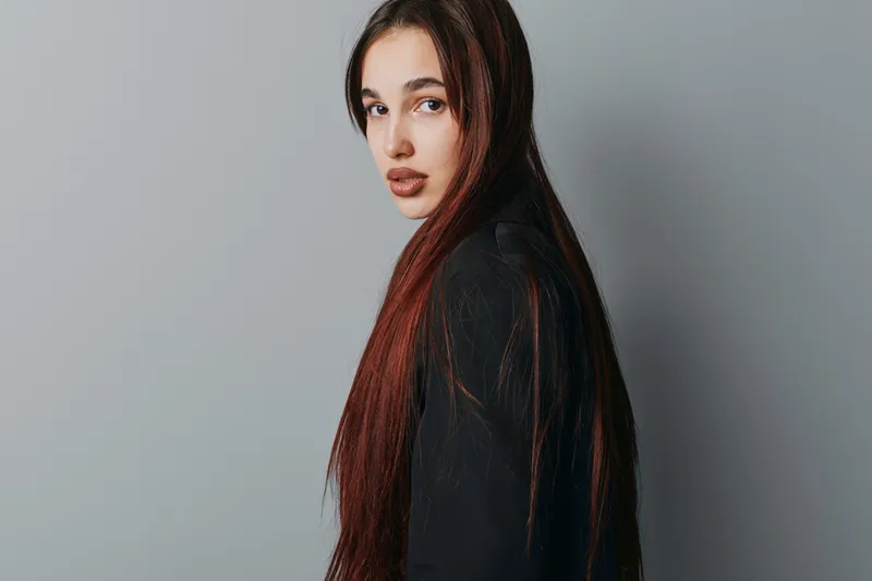 Young woman with long brown hair and natural makeup posing against gray background for beauty portrait, fashion style, and confident look with casual black clothing in studio.