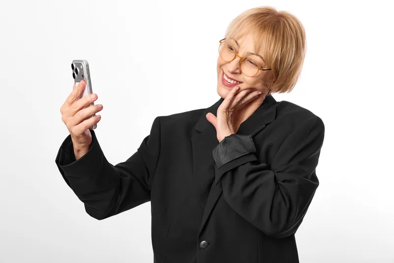 Elderly woman with glasses takes a selfie using a phone, smiling toward camera in a studio portrait, white background, professional jacket and coat, soft lighting, indoor calm