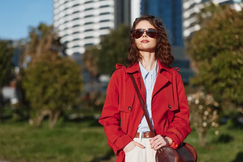 Stylish young woman with dark curly hair in a red trench coat stands in a sunny park, wearing sunglasses and a crossbody bag, posing confidently for a fashion shot.
