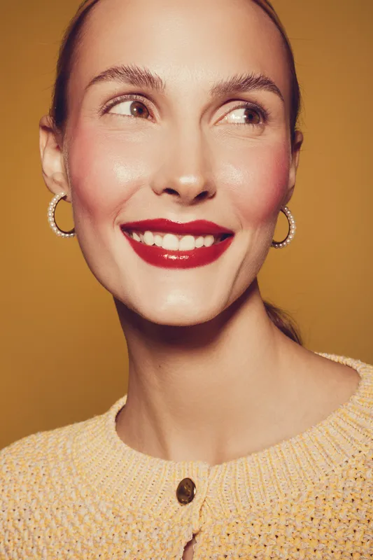 Smiling woman with red lipstick and hoop earrings looks to the side. She wears a yellow knitted sweater against a warm brown background. Concept of beauty, fashion, and happiness.