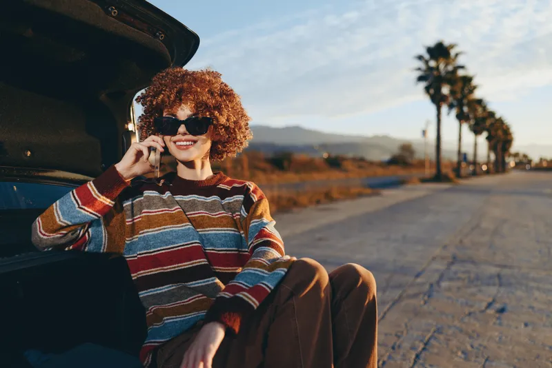 Woman lifestyle, curly hair, wearing sunglasses and striped sweater, sitting by open car door on an outdoor road, warm sunset light, palm trees and distant hills, calm relaxed