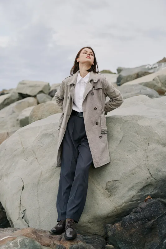 Young woman in long grey coat stands confidently on large rocks under cloudy sky. Fashion portrait of stylish model in casual outfit outdoors with natural background and moody atmosphere.
