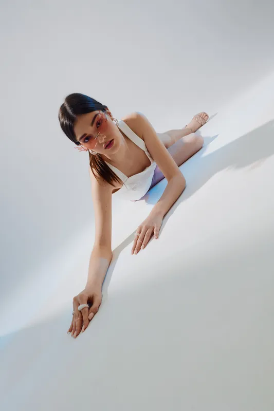 Young woman in white fashion outfit poses on a white surface with stylish sunglasses. She looks directly at the camera with a confident expression. Concept of beauty and fashion.