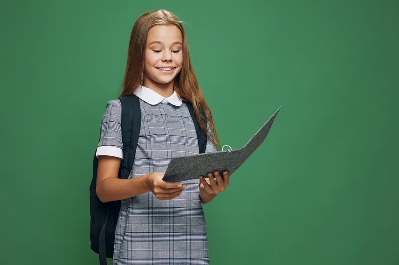 Schoolgirl student with backpack reading book or notebook with smile on green background for education, learning, knowledge and study at school or academy.
