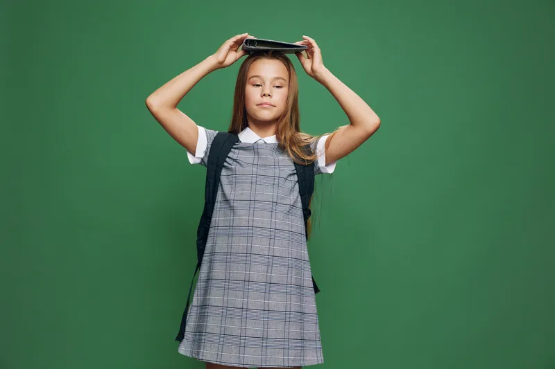 Girl with closed eyes standing against green background holding book on head wearing checkered dress and backpack for school or education concept in studio.