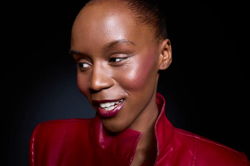 Close-up portrait of a smiling black woman with dark skin wearing a red leather jacket against a black background. Concept for fashion, beauty, confidence, and style in studio lighting.