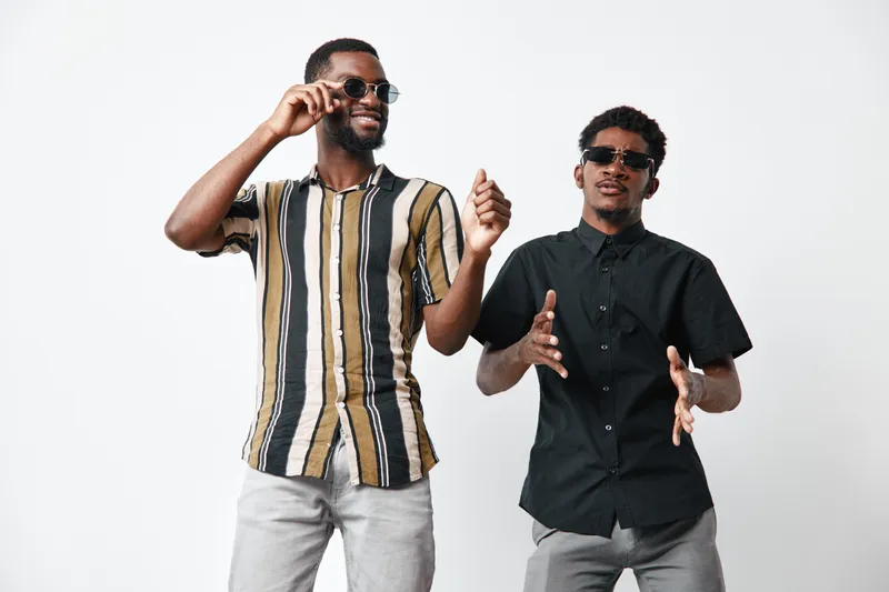 Two African American men with dark skin wearing sunglasses and casual shirts dancing and enjoying music together on white background in studio. Friends having fun and smiling.