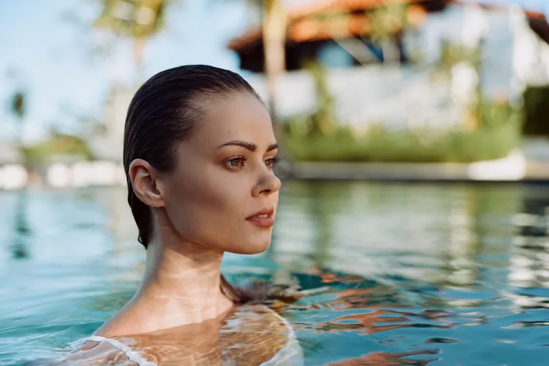 Relaxed woman floats in clear pool water, close up portrait with serene expression, sunlit resort surroundings, lifestyle leisure moment by the water