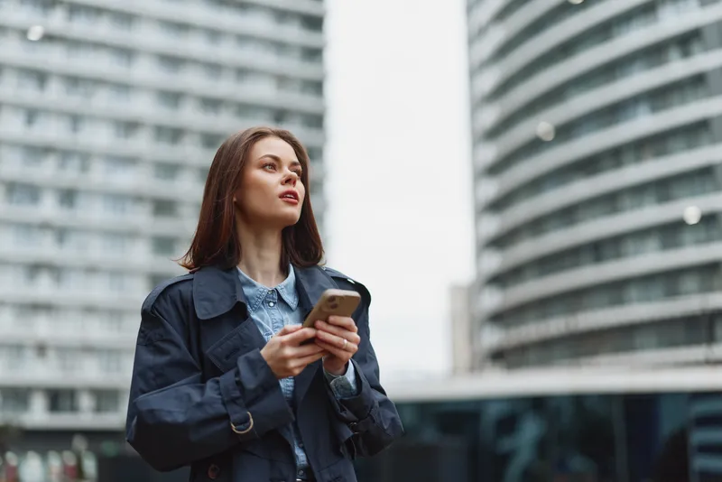 Young woman in blue coat holds smartphone looks away with confident expression standing in modern city with tall glass buildings background.
