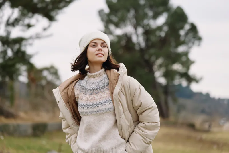 Woman in outdoor winter scene wearing a cozy sweater and beige jacket with a knit beanie, standing in a calm landscape with distant trees and a soft sky