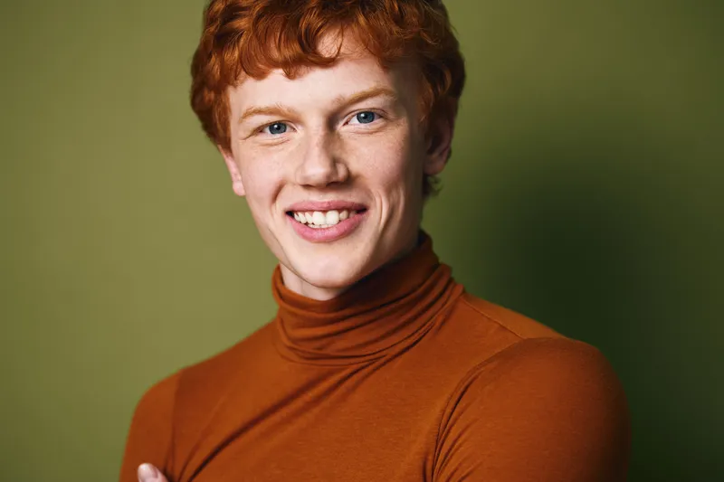 Portrait of a young man with red hair smiling wearing a brown turtleneck sweater against green background, showing confidence and friendly expression indoors.