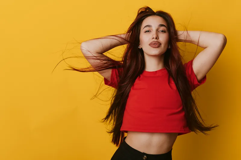 Young woman with long hair wearing red cropped top posing with hands behind head against yellow background. Casual style, confident expression and natural beauty in studio portrait.
